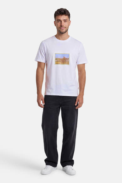 T-shirt blanc de la marque ORIGINALS BY JACK & JONES avec motif timbre paysage et texte, porté avec un jean foncé et des baskets blanches.