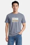 T-shirt gris-bleu à manches courtes de ORIGINALS BY JACK & JONES avec imprimé timbre "Offline." et scène de plage, porté par un homme en jeans.