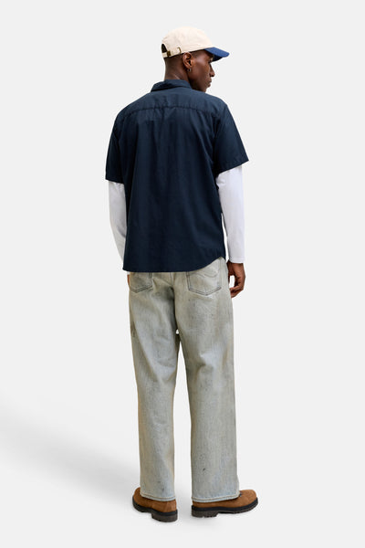 Chemise bleue à manches courtes de JACK & JONES, portée sur un t-shirt blanc à manches longues, avec un pantalon délavé et des chaussures marron.