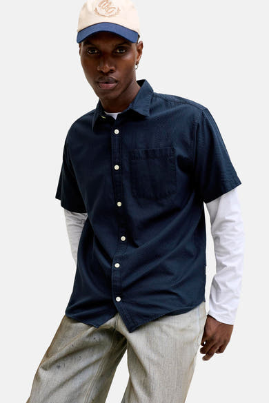 Blauw overhemd met korte mouwen van JACK & JONES, gedragen over een T-shirt met lange mouwen en een verbleekte spijkerbroek, met een pet.