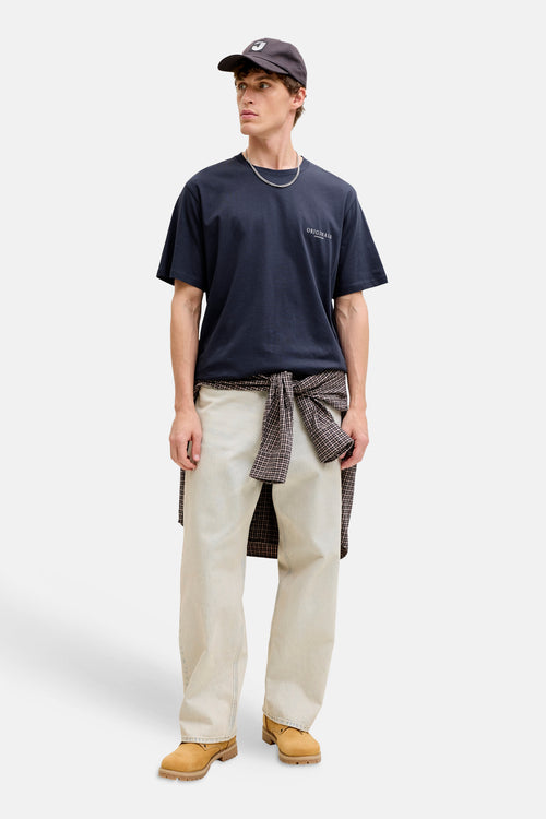 T-shirt bleu à manches courtes par ORIGINALS BY JACK & JONES, avec inscription, porté avec un pantalon clair et une chemise à carreaux nouée.