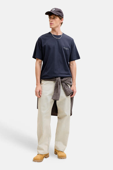 T-shirt bleu à manches courtes par ORIGINALS BY JACK & JONES, avec inscription, porté avec un pantalon clair et une chemise à carreaux nouée.