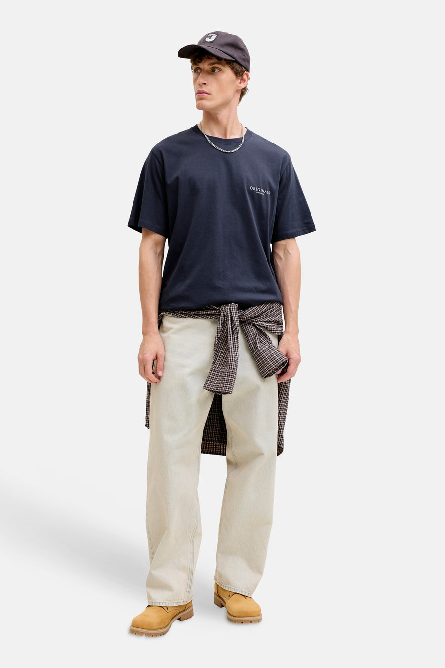 T-shirt bleu à manches courtes par ORIGINALS BY JACK & JONES, avec inscription, porté avec un pantalon clair et une chemise à carreaux nouée.