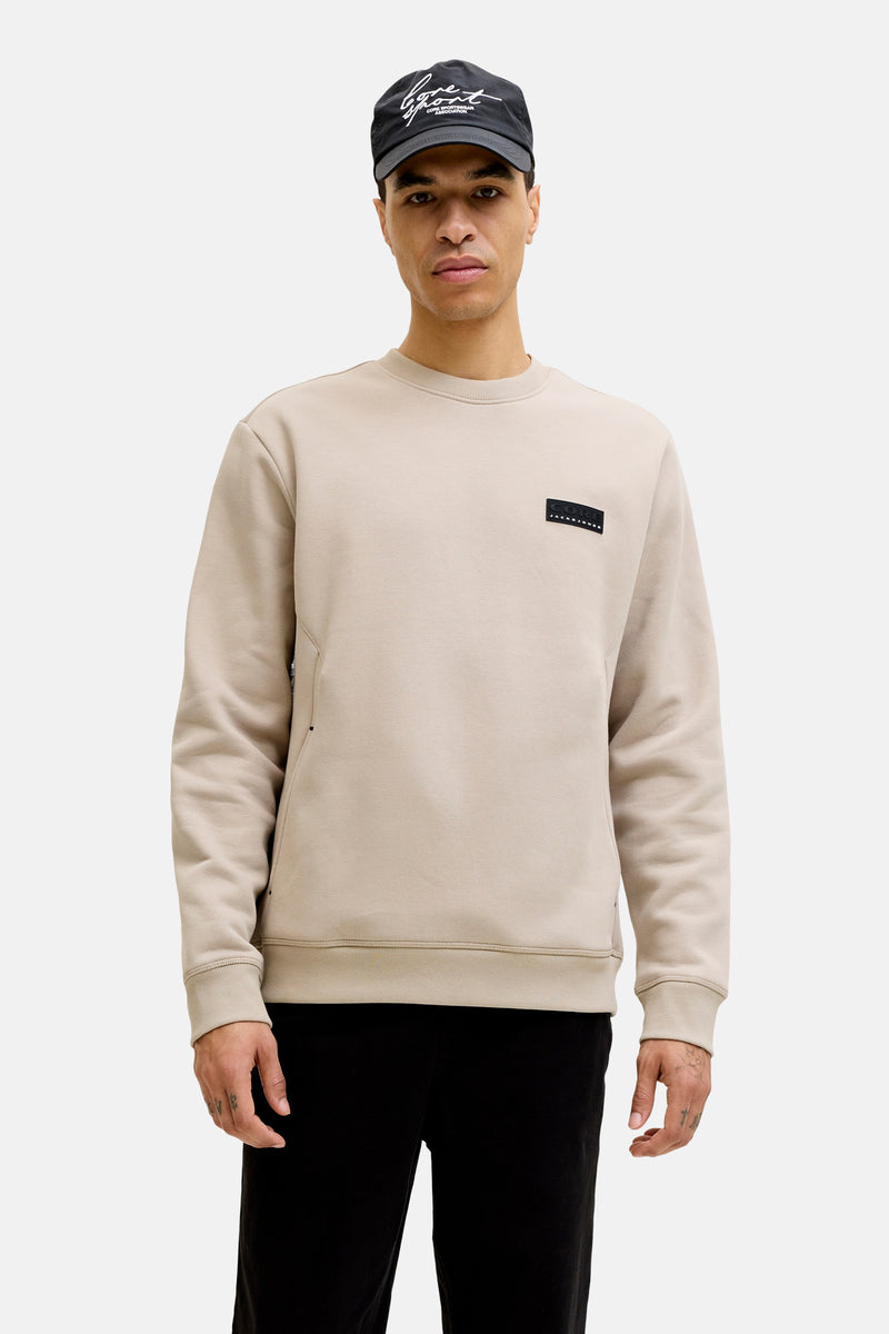 Beige ronde hals sweatshirt van Jack & Jones, gedragen met een zwarte gewatteerde jas en een zwarte joggingbroek.