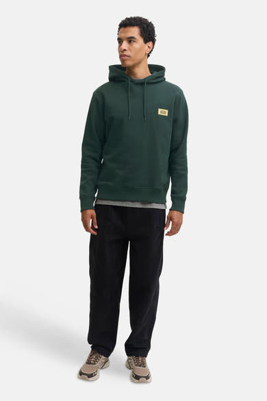 Dark green hooded sweatshirt with front patch logo by Jack & Jones, worn with black pants and tan sneakers, full-length front view