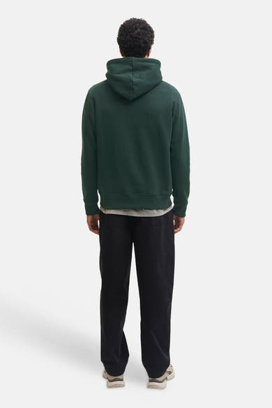 Dark green hooded sweatshirt by Scarab, back view, ribbed cuffs and hem, worn with black pants and beige sneakers.
