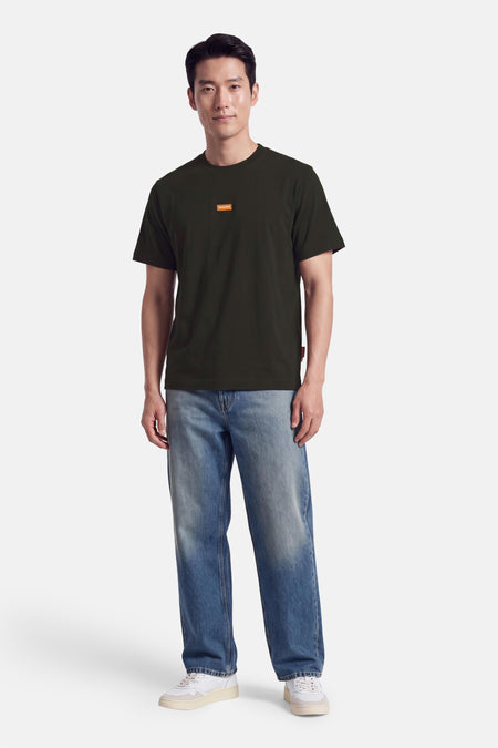 T-shirt vert à manches courtes de JACK & JONES, col rond, logo orange sur poitrine, porté avec jeans délavés et baskets blanches, vue de face.