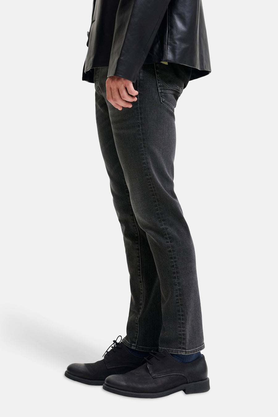 Black slim-fit denim jeans by Glenn, side view showing textured fabric, clean stitching, and tapered leg with black leather jacket and shoes.