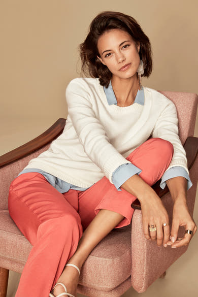 Pull blanc à col rond de VILA, porté avec un chemisier bleu et un pantalon corail, assise dans un fauteuil.