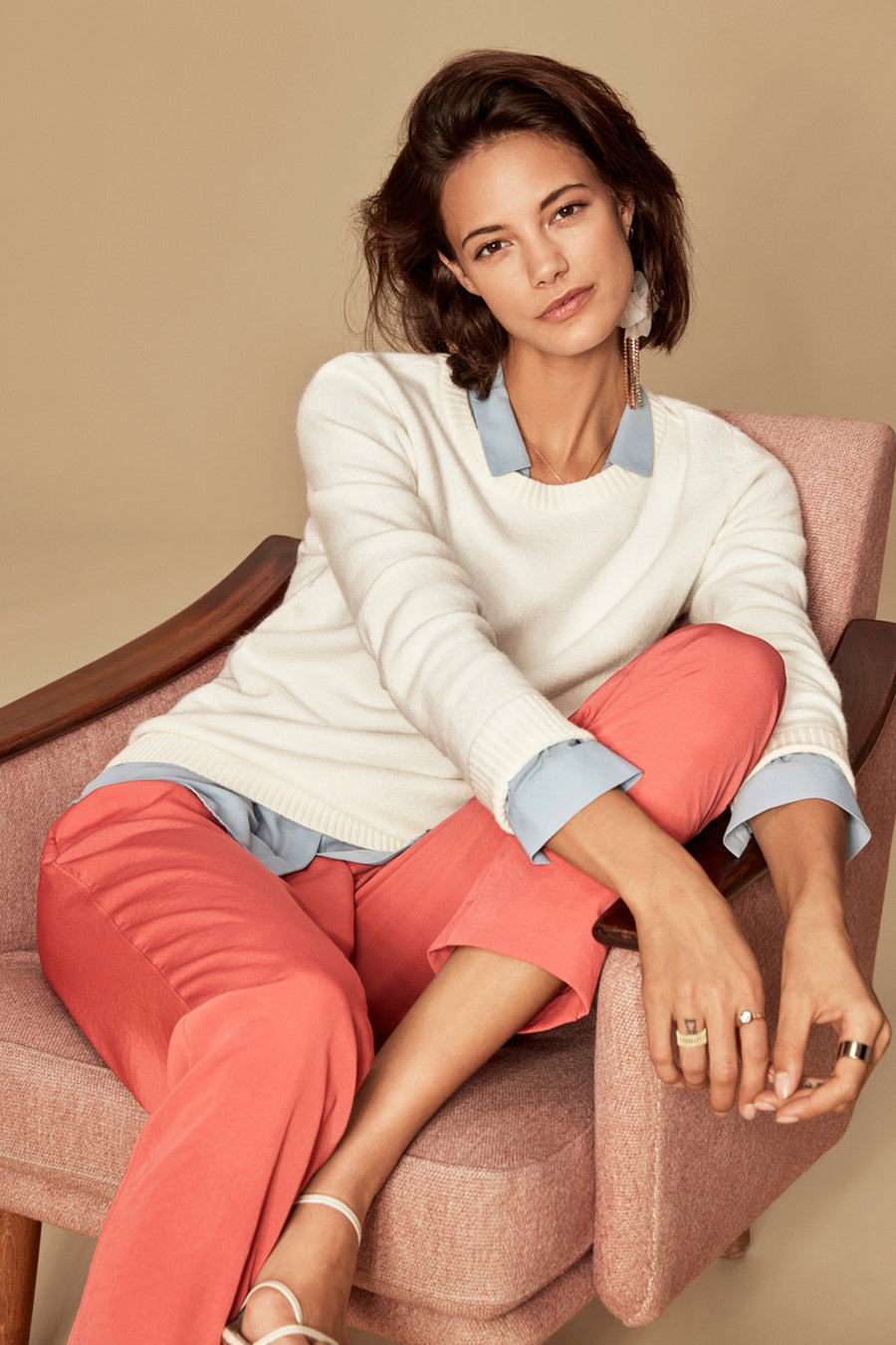 Pull blanc à col rond de VILA, porté avec un chemisier bleu et un pantalon corail, assise dans un fauteuil.