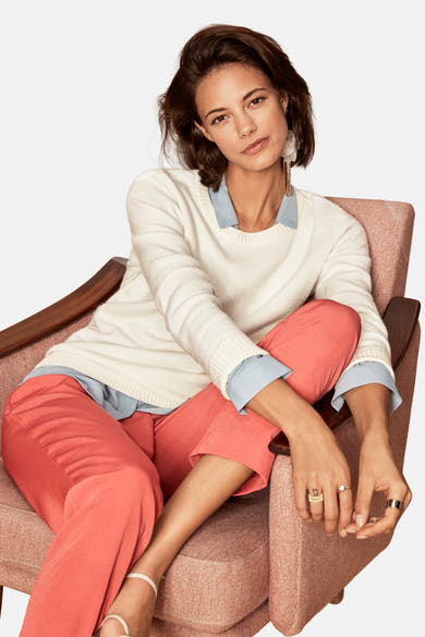 Pull col O blanc de VILA, porté avec une chemise bleu clair et un pantalon corail, assis dans un fauteuil.