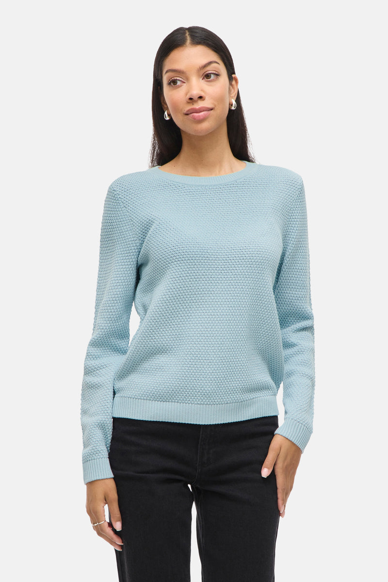 Pull bleu à col rond par VILA, texture tricotée, porté avec un pantalon noir.
