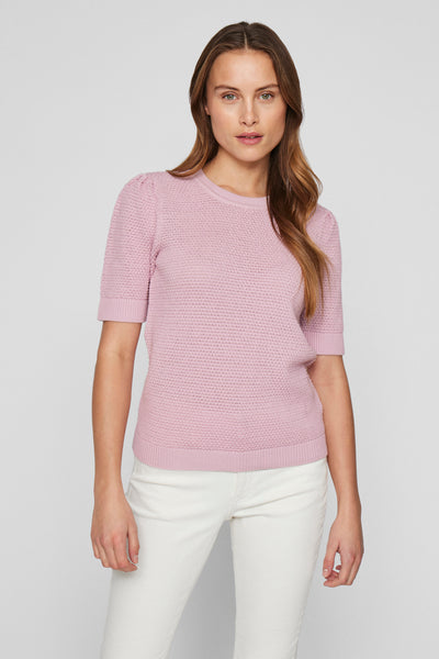 Pull rose en maille de VILA, avec manches courtes bouffantes, porté avec un pantalon blanc.