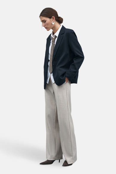 Beige VILA® broek, wijdvallend met een subtiele textuur, gedragen met een marineblauw colbert, een wit overhemd en een bruine stropdas.