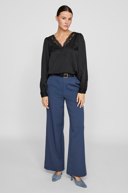 Blauwe broek van VILA, uitlopend model, gedragen met een zwarte riem en een zwarte blouse met kant.