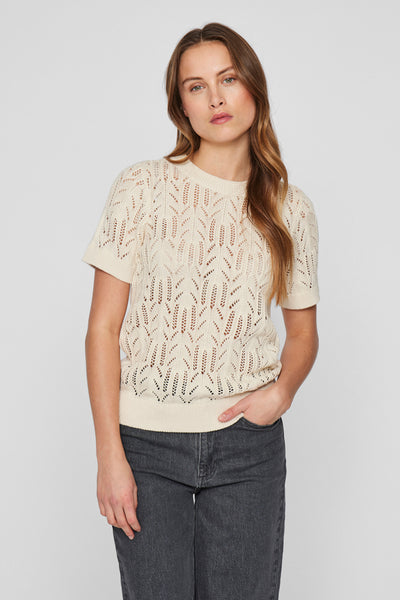 Pull à col rond écru de VILA®, avec motif ajouré et manches courtes, porté avec un jean gris.
