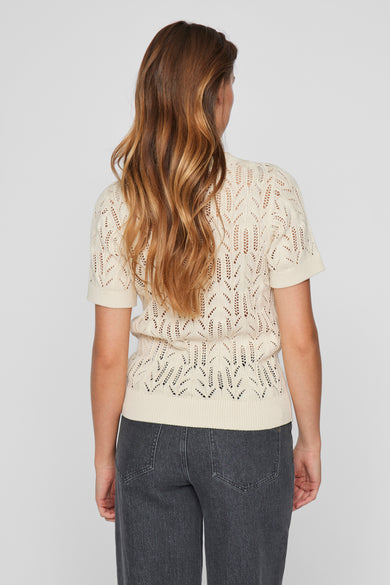 Pull écru à col rond de VILA®, en maille ajourée avec des motifs délicats, porté avec un jean gris.
