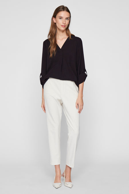 Blouse noire à manches longues de VILA, avec des poignets boutonnés, portée avec un pantalon blanc et des escarpins.
