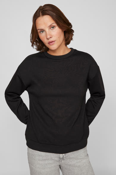 Zwarte sweater van VILA, gedragen met een lichtgrijze broek, handen achter de rug.