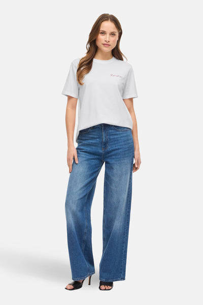 T-shirt blanc à manches courtes de VILA®, avec l'inscription 'Amore', porté avec un jean large et des sandales à talons.
