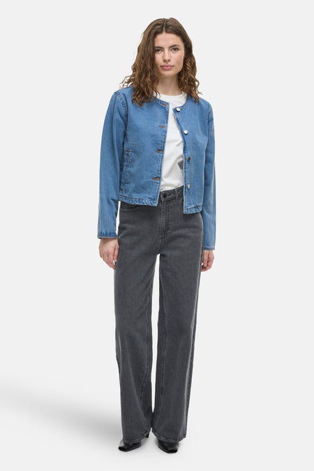 Kort jasje van middieblauw denim van VILA, gedragen over een wit T-shirt en een grijze wijde broek.