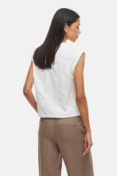Wit mouwloos topje van VILA®, met reliëf bloemenpatroon, gedragen met een bruine broek.
