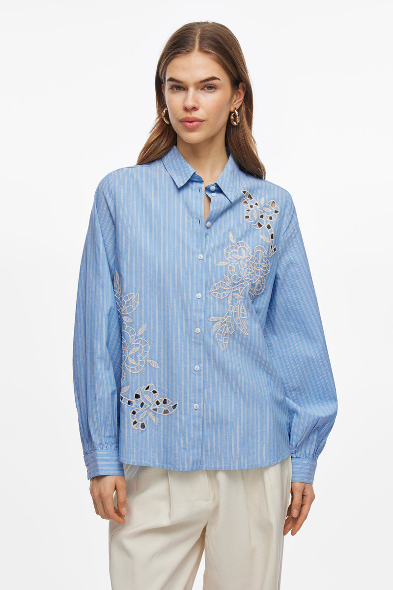 Blauwe blouse met lange mouwen van VILA®, versierd met witte bloemenborduursels, gedragen met een beige broek en bijpassende pumps.
