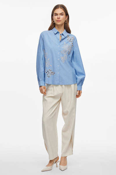 Chemisier bleu à manches longues de VILA®, orné de broderies florales blanches, porté avec un pantalon beige et des escarpins assortis.
