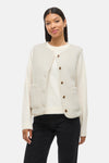 Gilet écru par VILA®, avec une texture bouclée, poches plaquées et boutons marron, porté sur un pull et un jean noir.