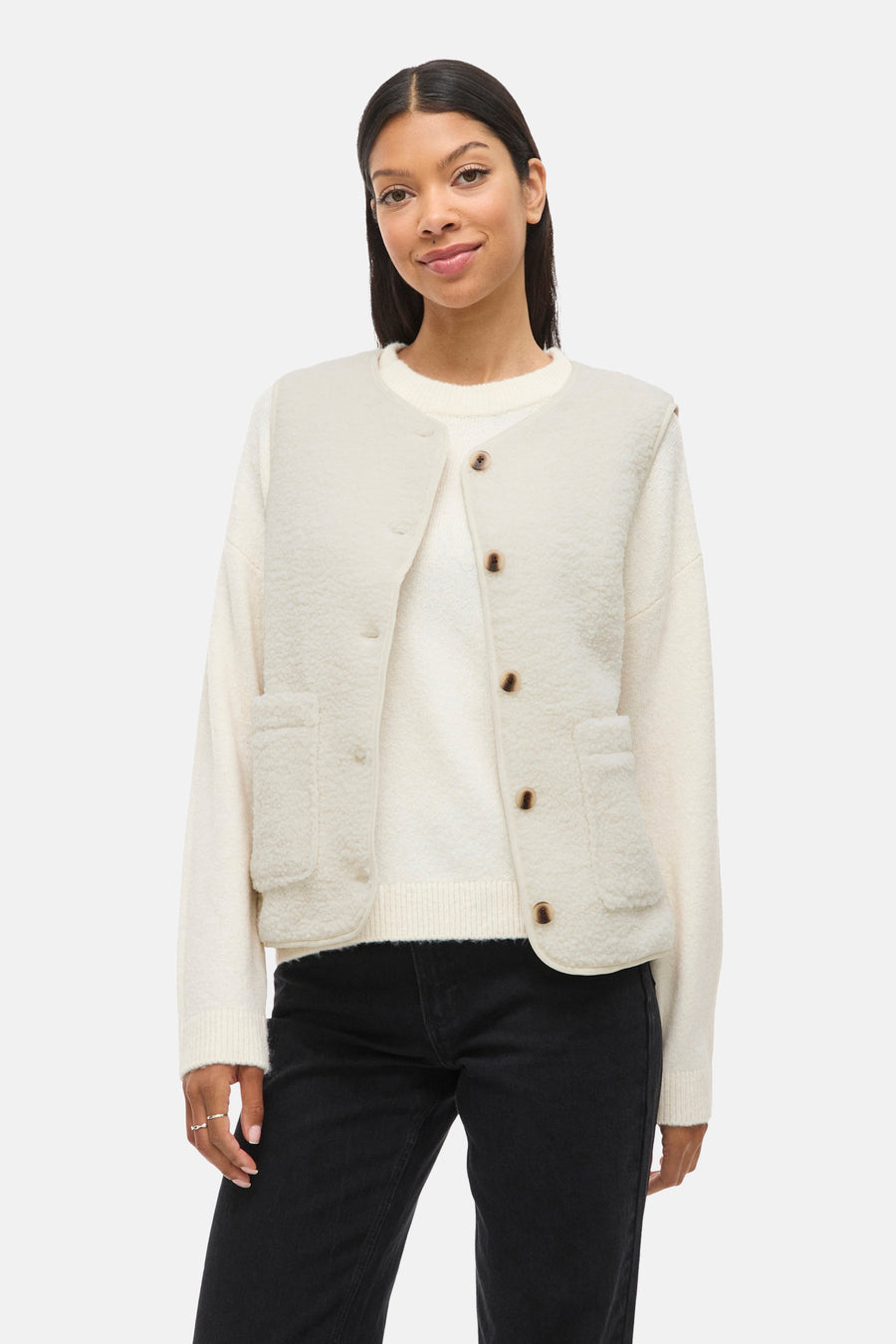 Gilet écru par VILA®, avec une texture bouclée, poches plaquées et boutons marron, porté sur un pull et un jean noir.