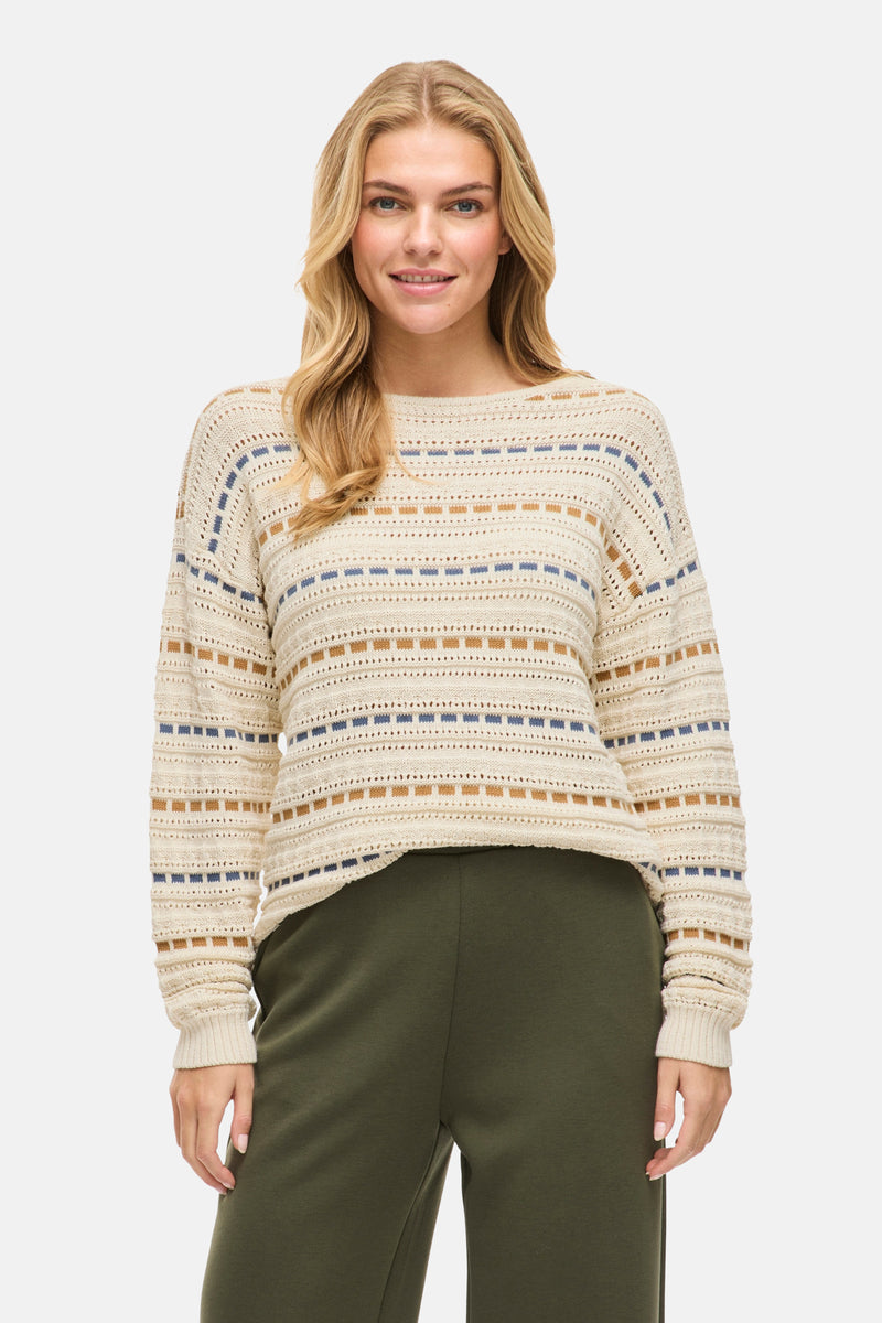 Pull à col rond beige de VILA®, avec motifs géométriques bleus et marrons, porté avec un pantalon vert.
