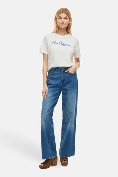 T-shirt à manches courtes blanc de VILA®, avec l'inscription bleue « Ciao Ragazzi », porté avec un jean et des chaussures marron.
