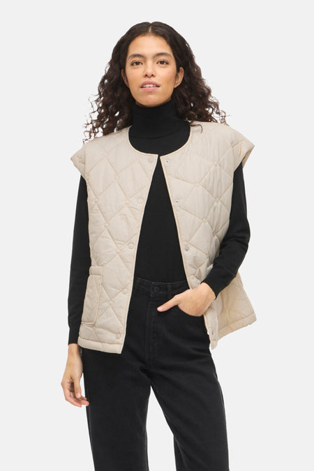 Veste sans manches beige de VILA®, à motif matelassé et poches plaquées, portée avec un pull noir et un jean.