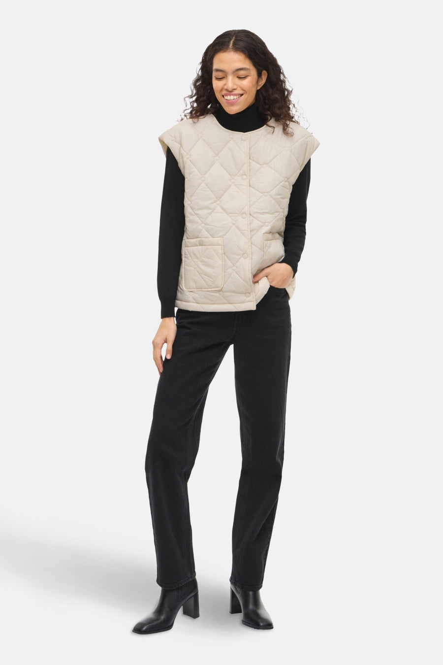 Beige mouwloos gewatteerd vest met ruitpatroon van VILA®, gedragen over een zwarte trui met een zwarte jeans.