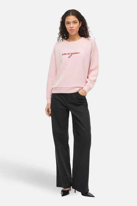Sweat-shirt rose de VILA®, avec le mot « imagine » en rouge, porté avec un pantalon noir.
