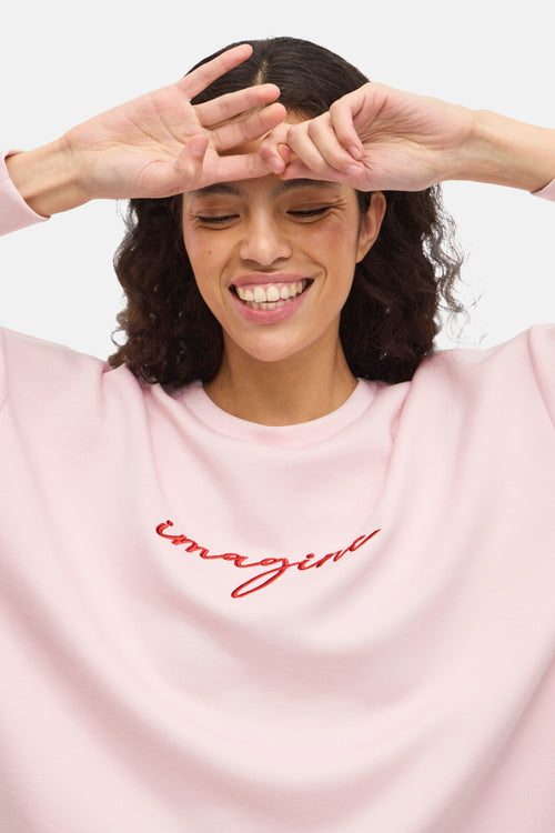 Sweat rose van VILA® avec l'inscription 'imagine' en rouge brodée sur le devant, porté par un mannequin souriant.
