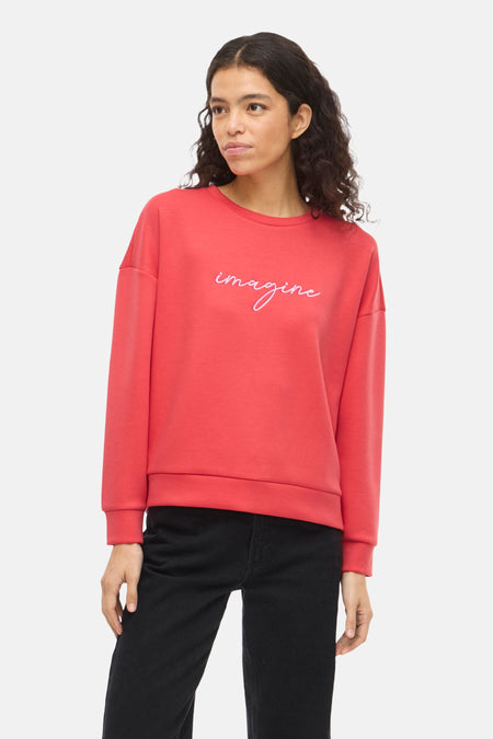 Oranje VILA® sweater met geborduurd "Imagine"-motief, gedragen met een zwarte jeans en zwarte naaldhakken.