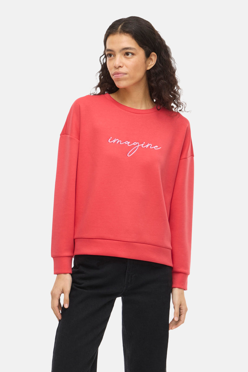 Oranje VILA® sweater met geborduurd "Imagine"-motief, gedragen met een zwarte jeans en zwarte naaldhakken.