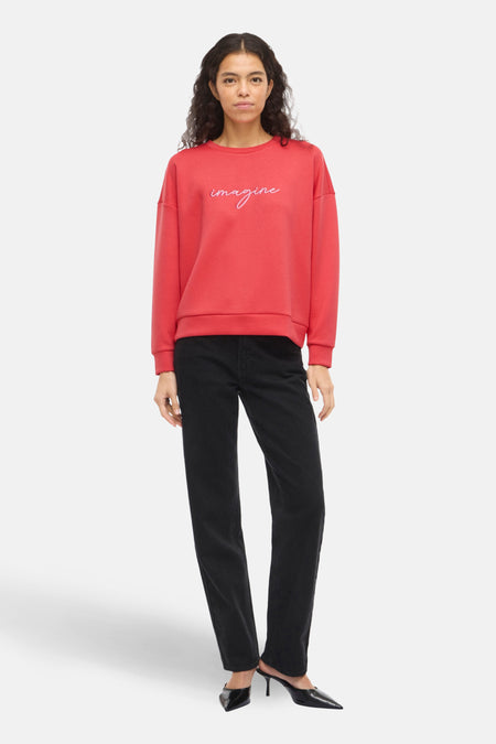 Sweat orange VILA® avec motif « Imagine » brodé, porté avec un jean noir et des escarpins noirs à talons aiguilles.