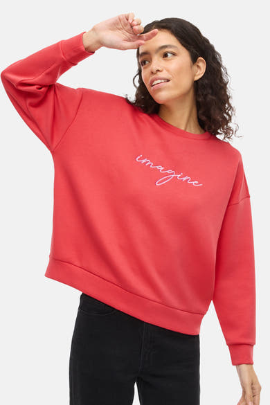 Oranje V-hals sweatshirt van VILA®, met roze geborduurde tekst "imagine", gedragen met een zwarte jeans.