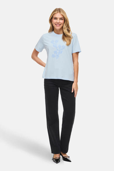T-shirt bleu à manches courtes de VILA, avec un motif floral imprimé, porté avec un jean noir et des escarpins.