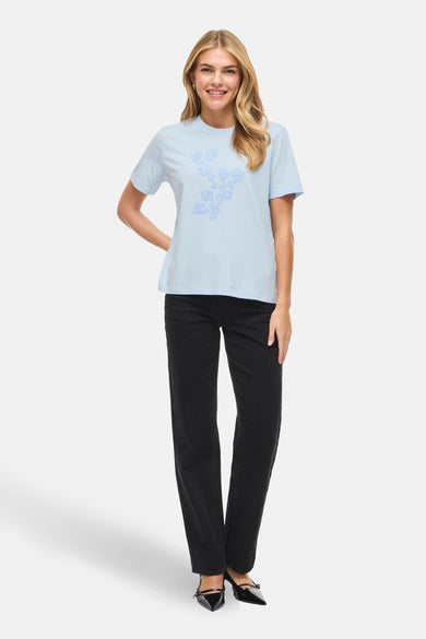 T-shirt bleu à manches courtes de VILA, avec un motif floral imprimé, porté avec un jean noir et des escarpins.
