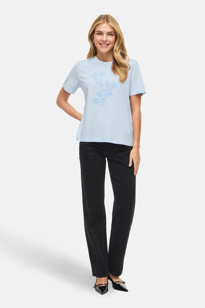 T-shirt bleu à manches courtes de VILA, avec un motif floral imprimé, porté avec un jean noir et des escarpins.