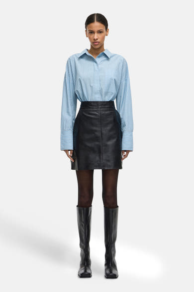 Chemise à manches longues bleue à rayures, de ROUGE EDIT, portée avec une jupe noire et des bottes hautes.