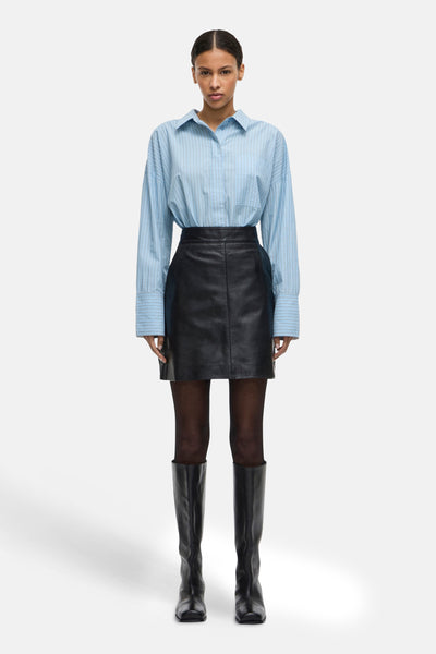 Chemise à manches longues bleue à rayures, de ROUGE EDIT, portée avec une jupe noire et des bottes hautes.