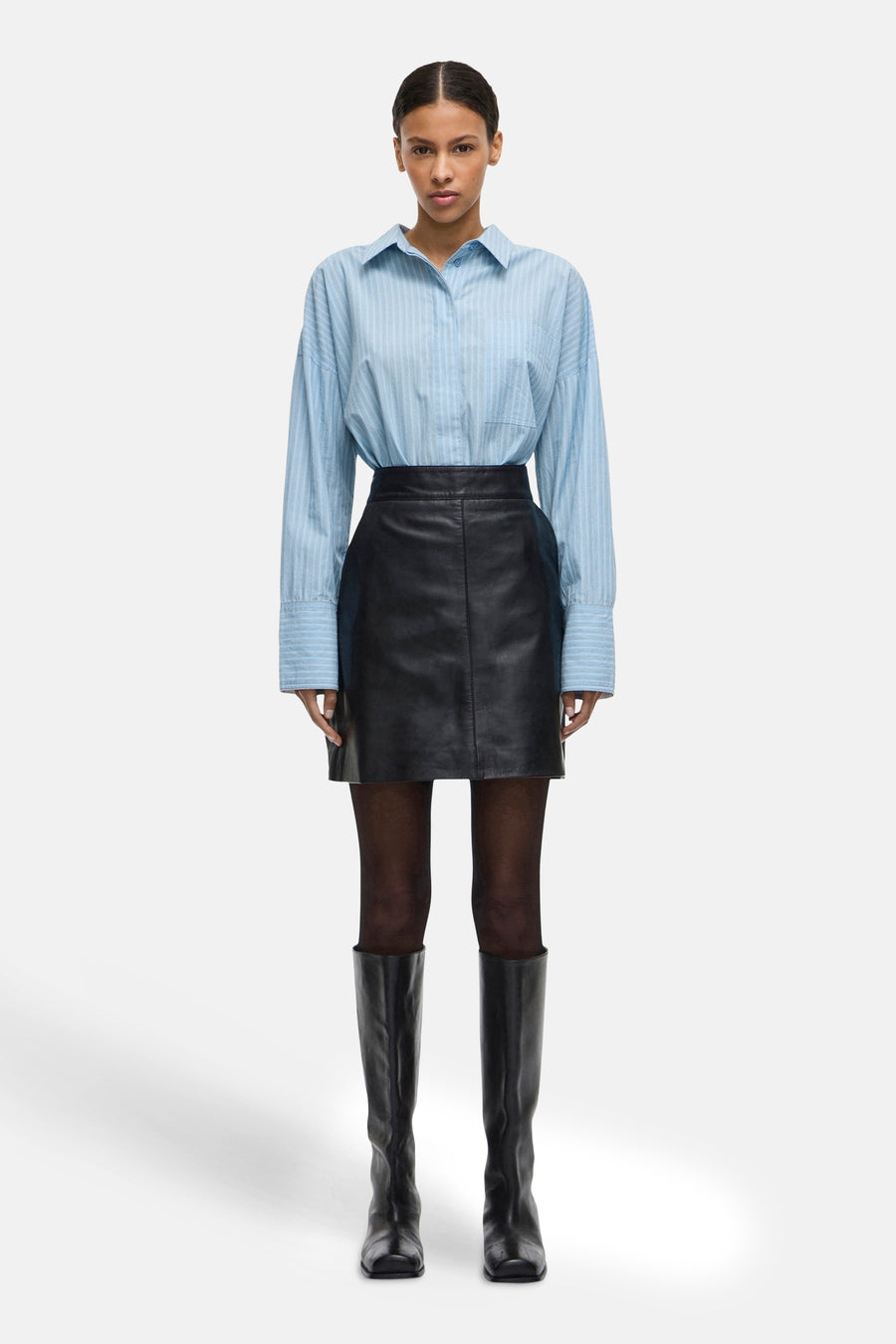 Chemise à manches longues bleue à rayures, de ROUGE EDIT, portée avec une jupe noire et des bottes hautes.