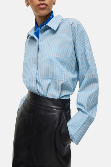 Chemise à manches longues bleu rayé de ROUGE EDIT, avec poche poitrine à logo, portée avec un foulard bleu et une jupe en cuir noir.