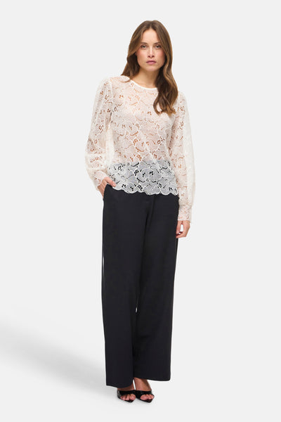 Blouse blanche à manches longues de VILA, en dentelle fleurie sur un fond clair, portée avec un pantalon noir.