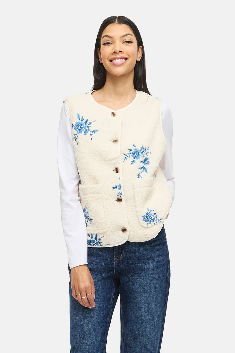 Beige mouwloos vest van VILA® met knopen en blauwe bloemenborduursel, gedragen over een witte top en een spijkerbroek.