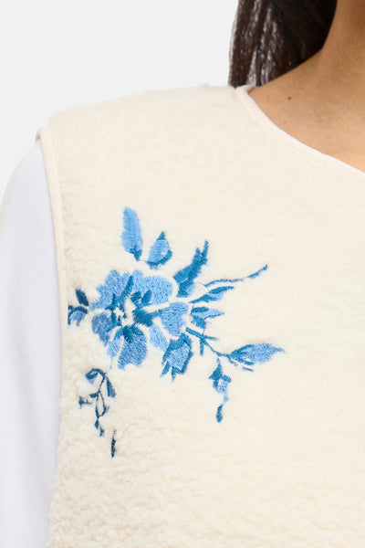 Beige mouwloos vest van VILA® met blauwe geborduurde bloemenprint op een pluizige stof.